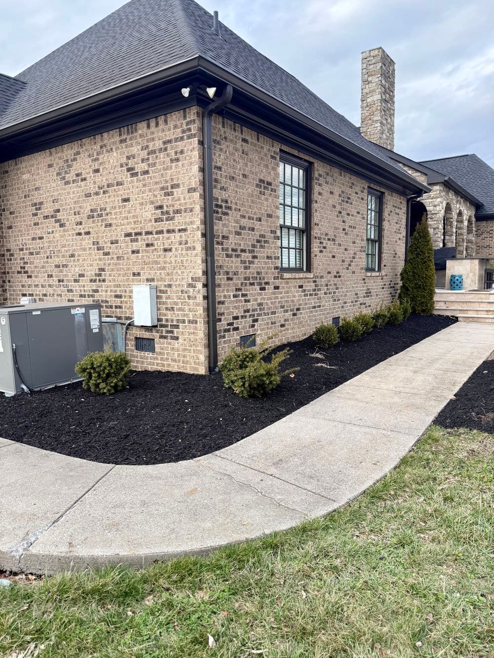 Side view of a brick house with shrubs, mulch, and air conditioning unit along the walkway.
